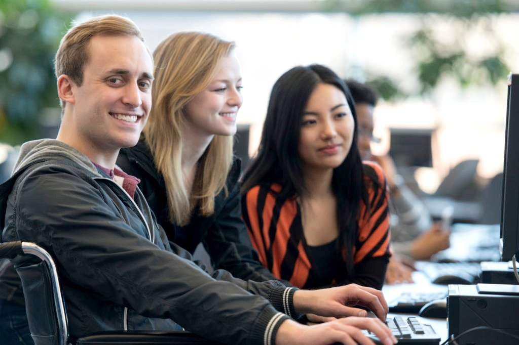 Persons With Disabilities - A diverse group of college age students are working on a group project in the computer lab. One man with a physical disability is sitting in a wheelchair and is smiling while looking at the camera.