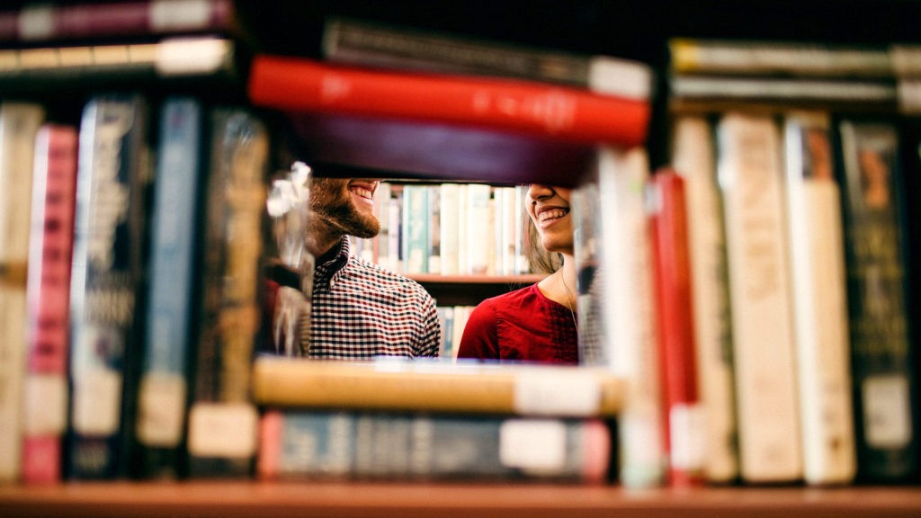 Learning Library - Two people smiling at each other, as seen through a frame of books in a library.